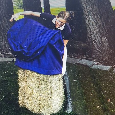 A person in equestrian gear stands outdoors, placing a blue protective cover over a rectangular hay bale.