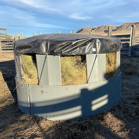 A round, grey plastic hay feeder with a black tarp cover sits in a dry, fenced livestock corral.