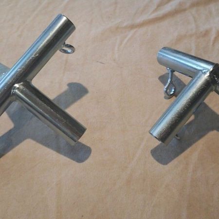 Two silver metal T-shaped and L-shaped connector pipes with small eyelet attachments rest on a beige fabric surface.