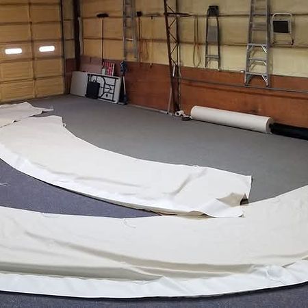 Two large, curved white fabric pieces laid out on a gray floor in a workshop with ladders and rolls of material behind.