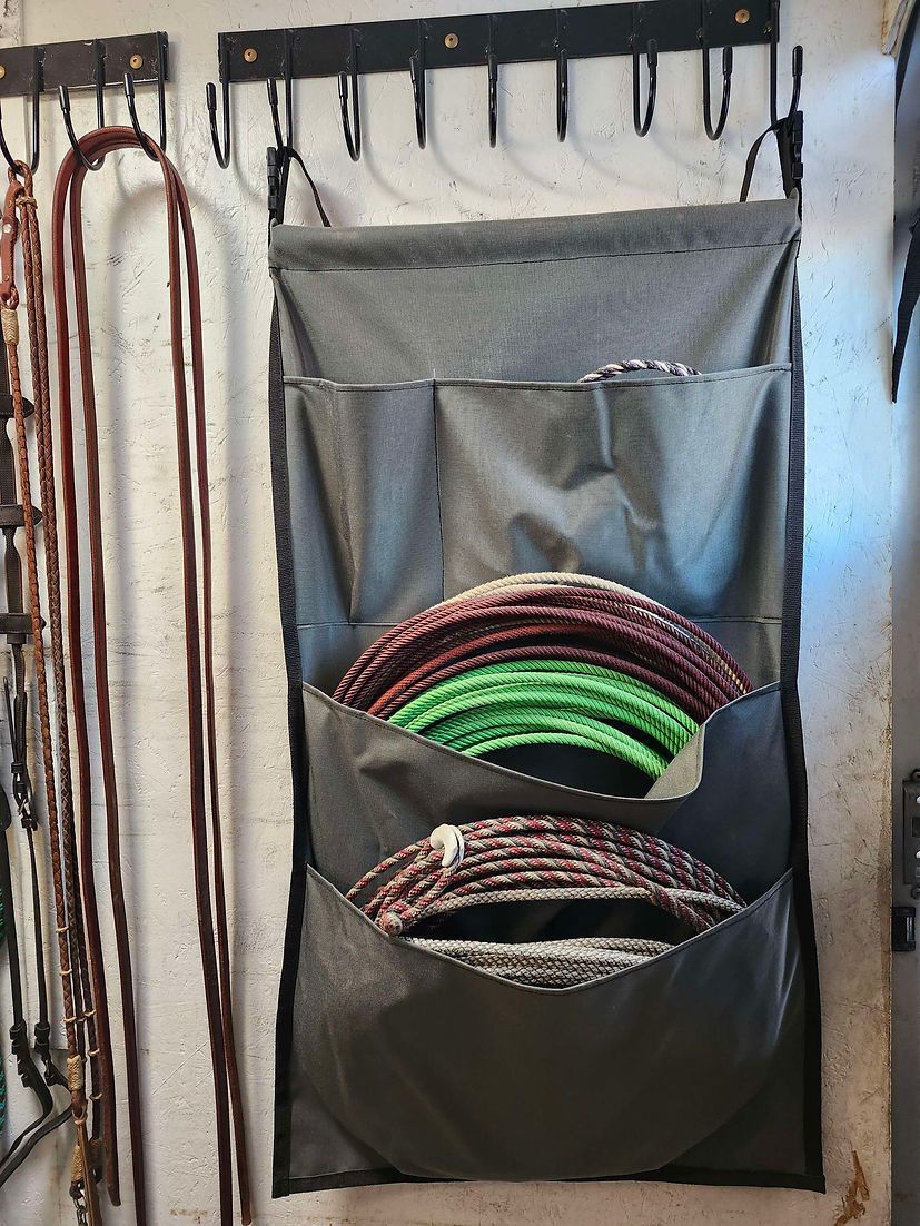 A wall-mounted hanging fabric organizer with pockets holding coiled ropes, next to a rack with hanging leather reins.