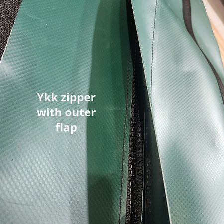 Close-up of a dark YKK zipper on a light green, textured fabric with an overlapping protective outer flap.