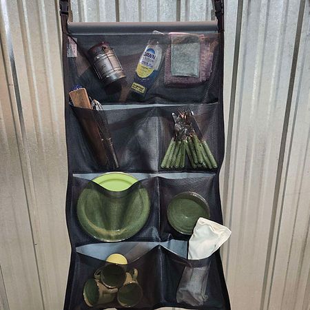 A black hanging organizer with four rows of pockets holding camping gear, including plates, utensils, and cleaning items.