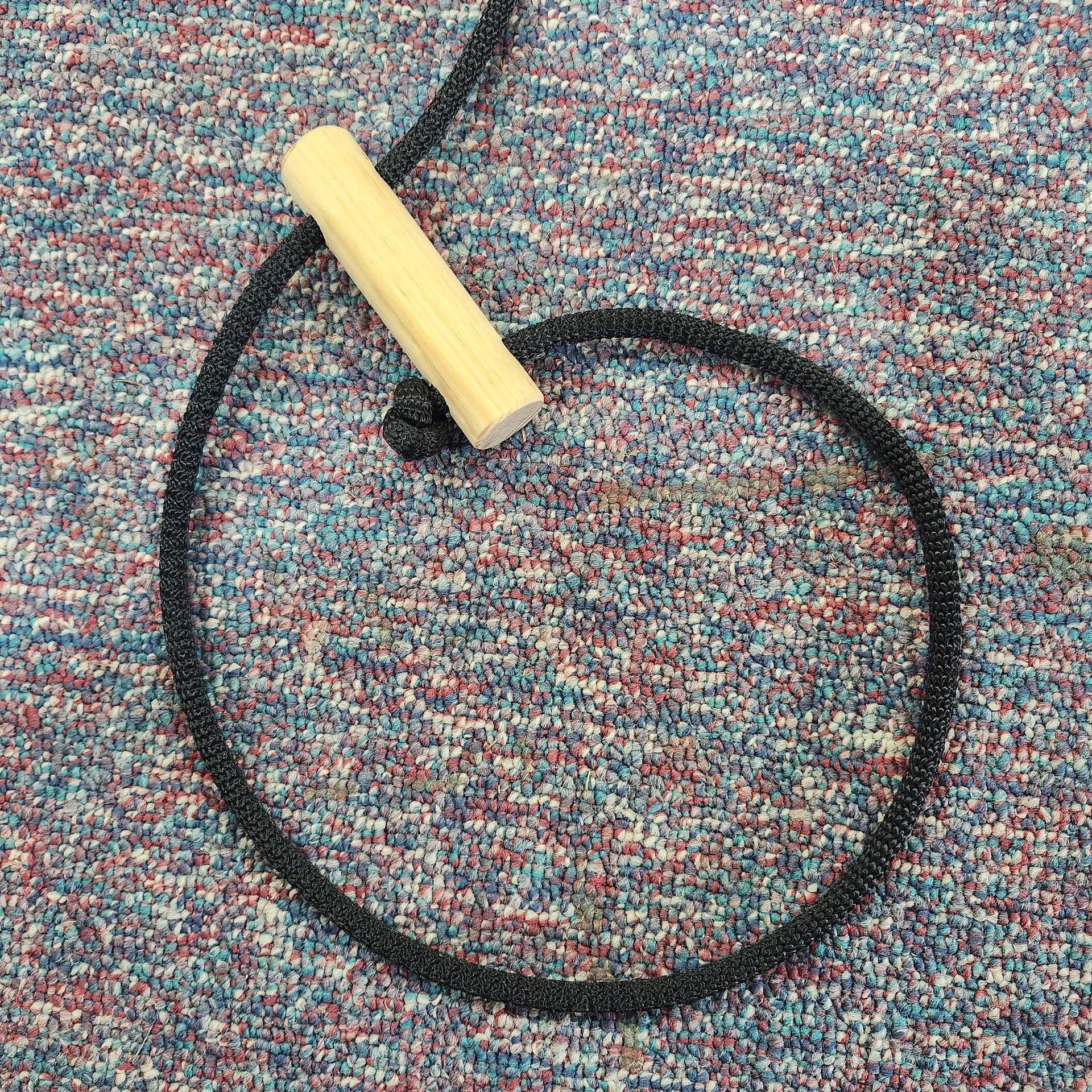 A single wooden handle attached to a black knotted rope lying in a circle on a multicolored speckled carpet.