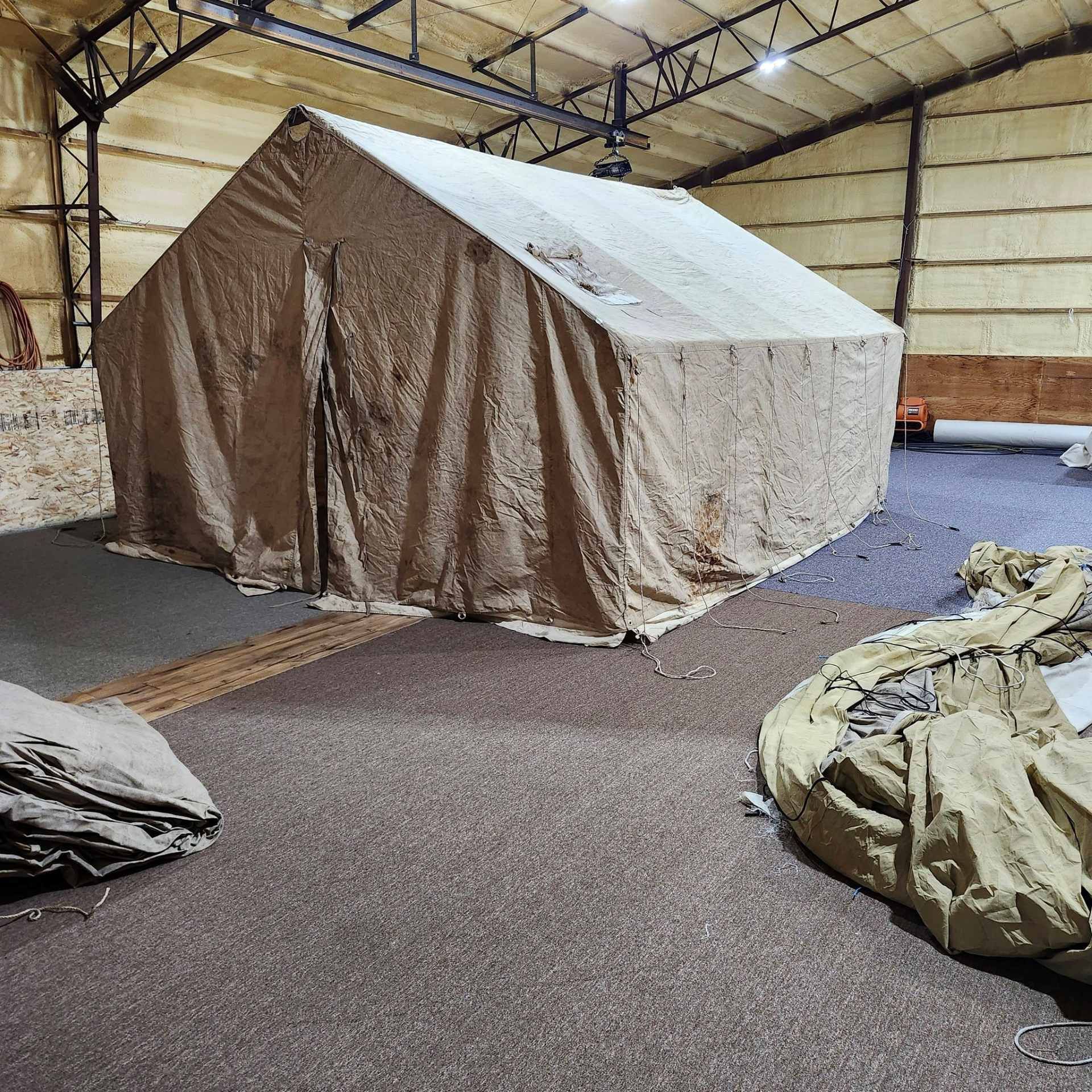 A tan, framed military-style shelter tent set up inside a large, industrial warehouse with concrete floors.