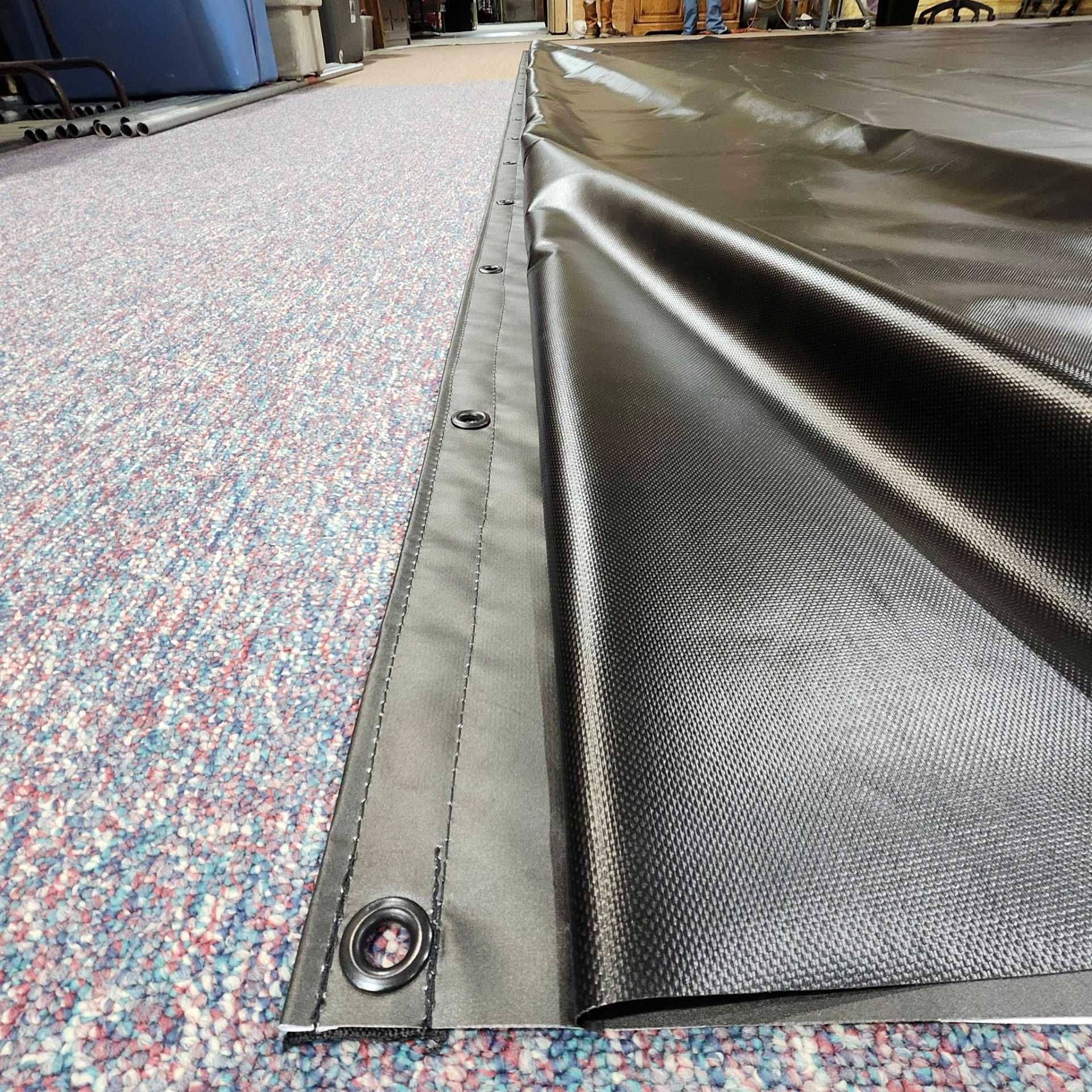 A close-up view of a black, heavy-duty tarp with metal grommets along a reinforced edge, lying on a speckled carpet.