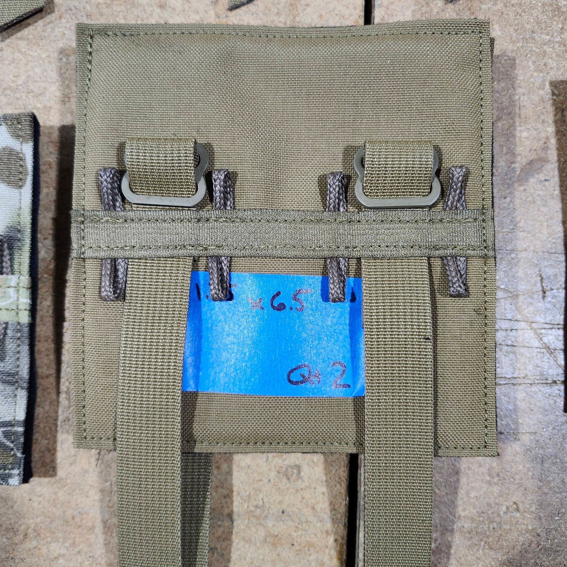 Tan tactical pouch back with mounting straps and plastic loops, featuring a blue label with handwritten text.