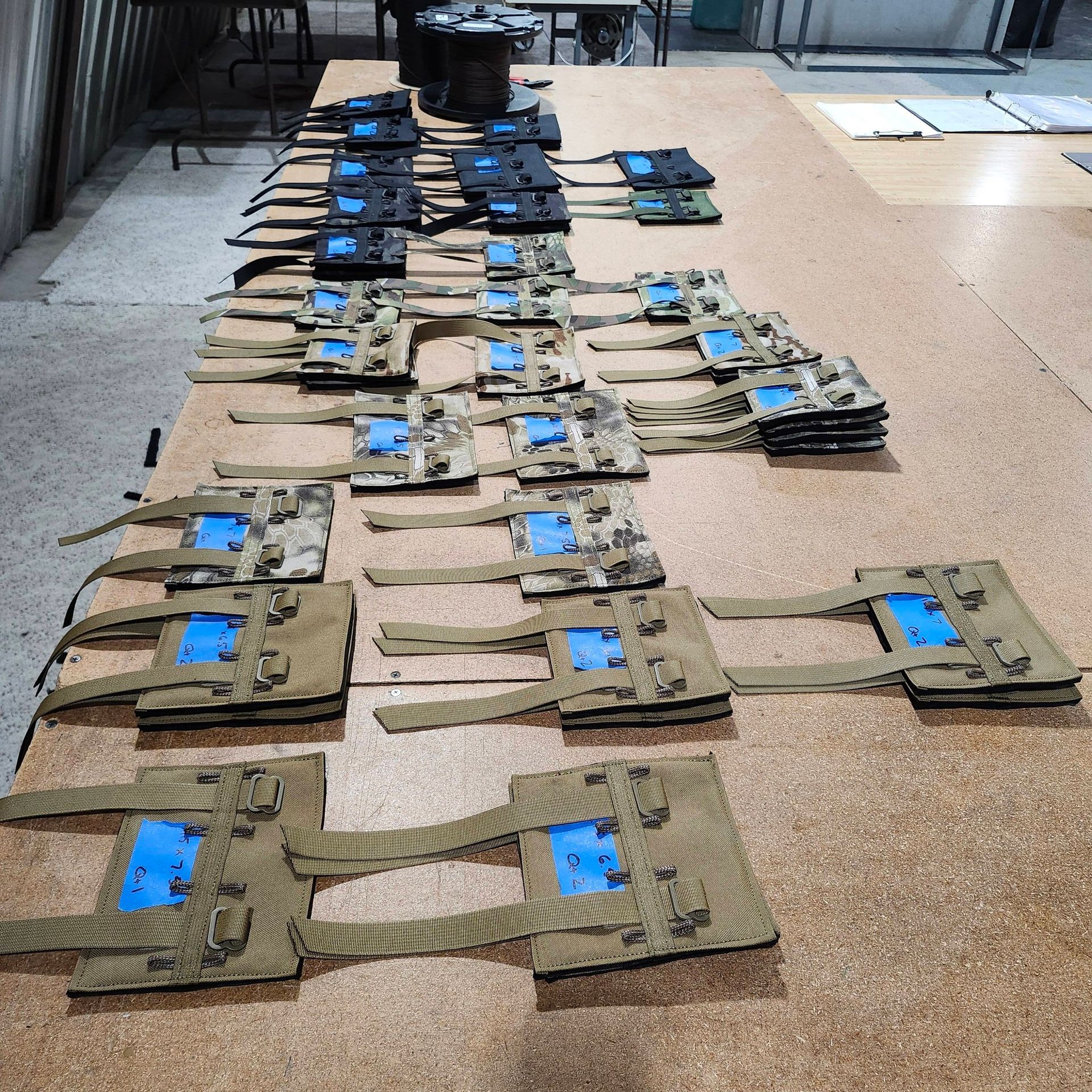 Rows of tactical vest components with blue identification tags arranged on a large table in a workshop setting.