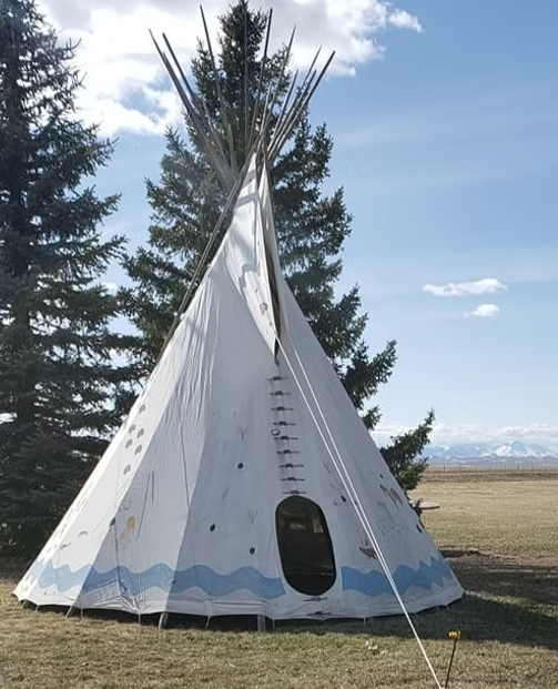 A white teepee with a blue wave pattern at the base stands outdoors near evergreen trees and a flat, grassy landscape.
