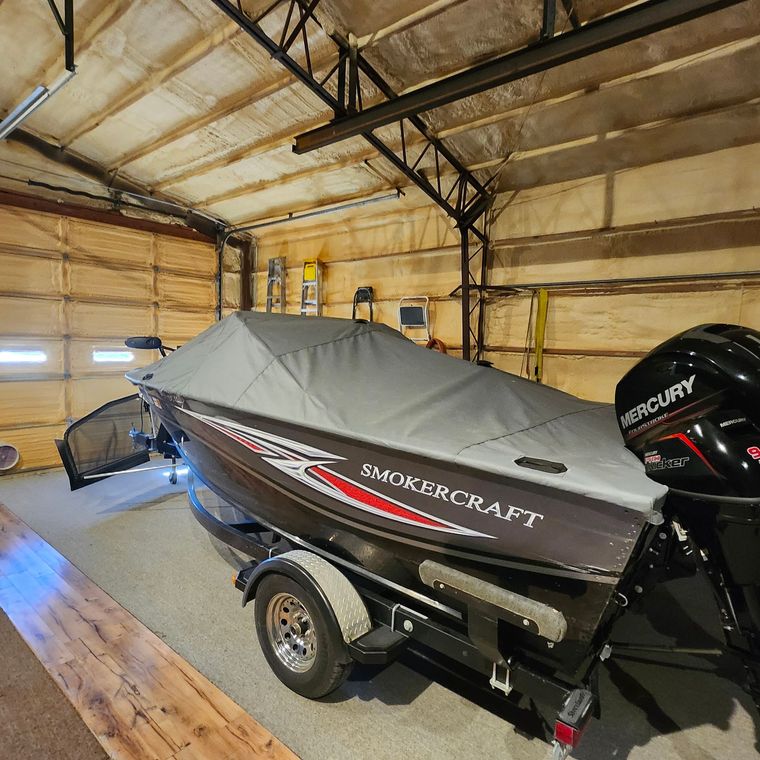 A SmokerCraft fishing boat with a gray cover on a trailer inside a workshop with wood-paneled walls and a Mercury motor.