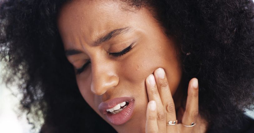 Woman with curly hair, eyes closed, touching cheek, grimacing in pain.
