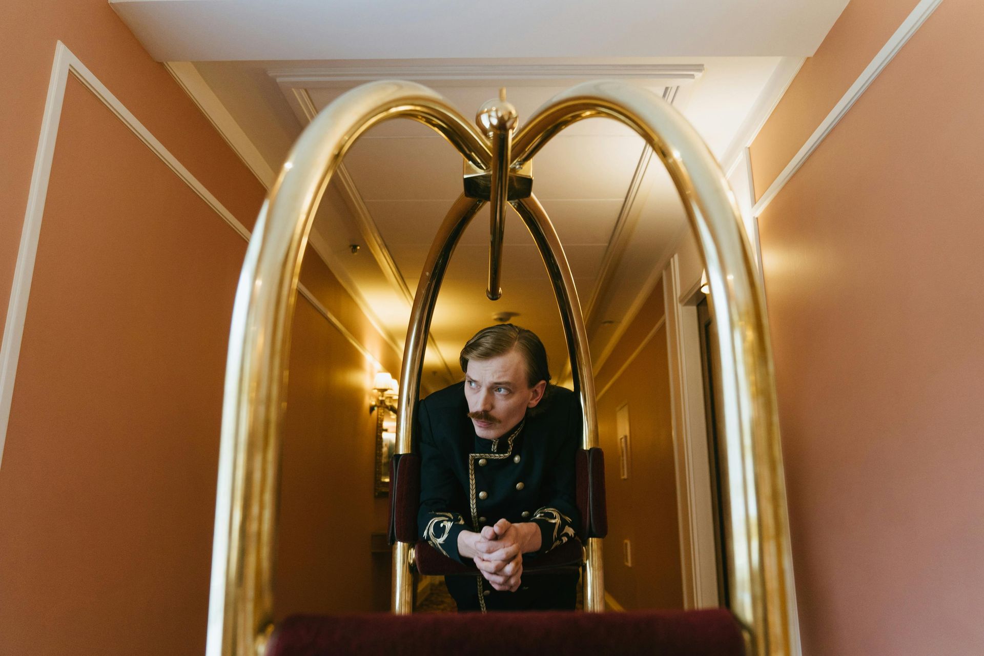 Bellhop in uniform stands behind a golden luggage cart in a hallway with peach walls.