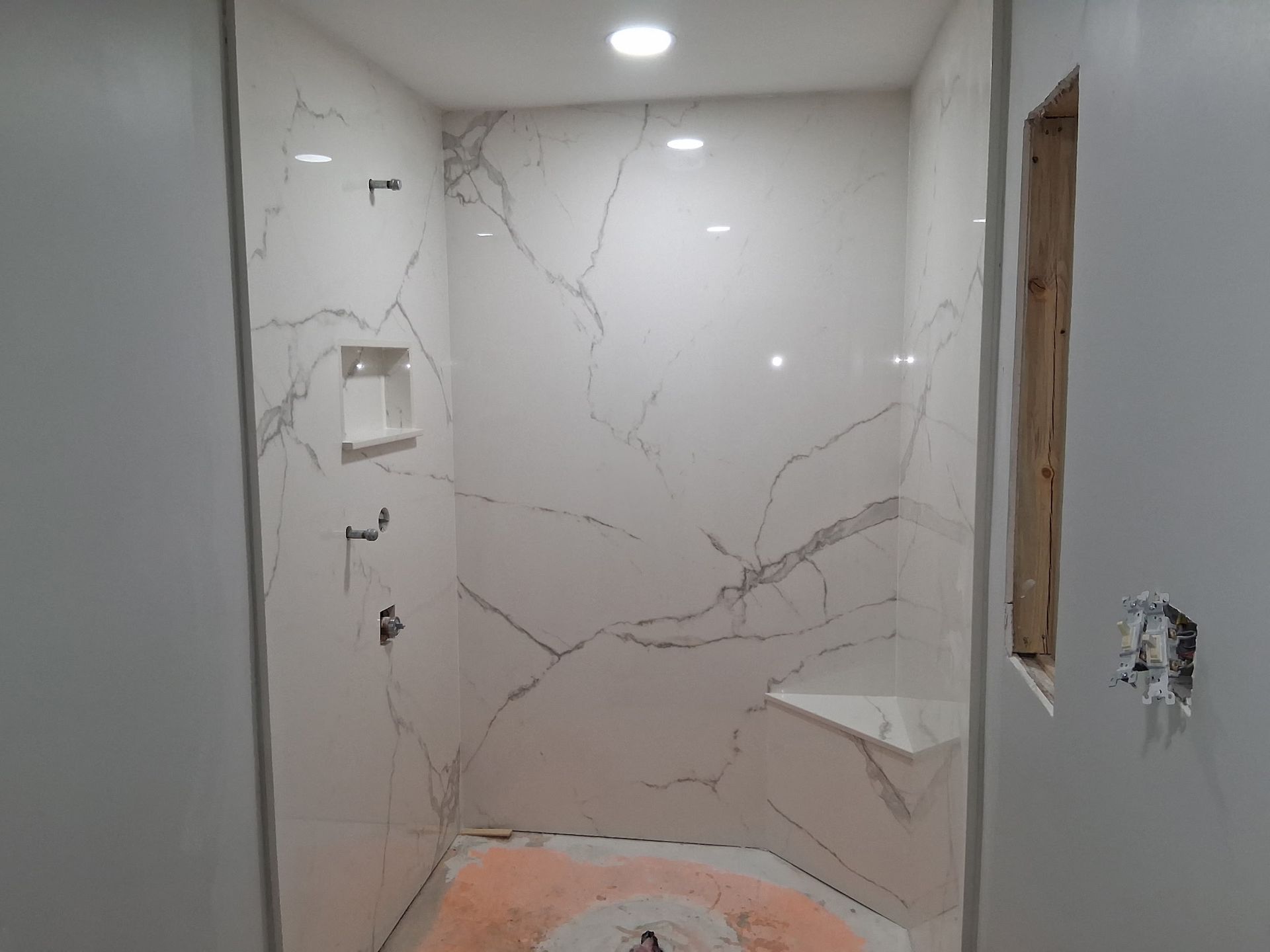 Bathroom shower under construction with white marble-look tile, recessed shelf, and a rough floor patch.