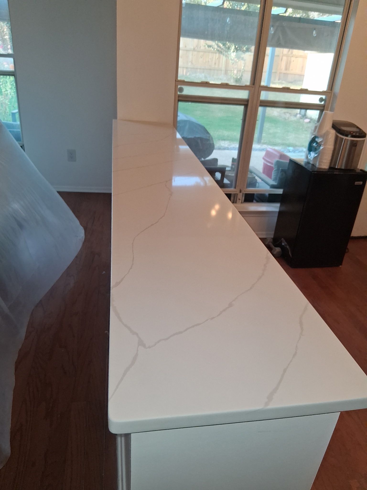Long white kitchen island with marble-like countertop in a bright room near large windows.