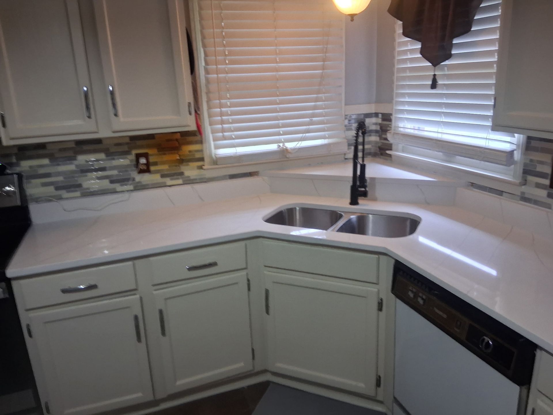 Modern kitchen corner with white cabinets, light countertops, double sink, and window blinds