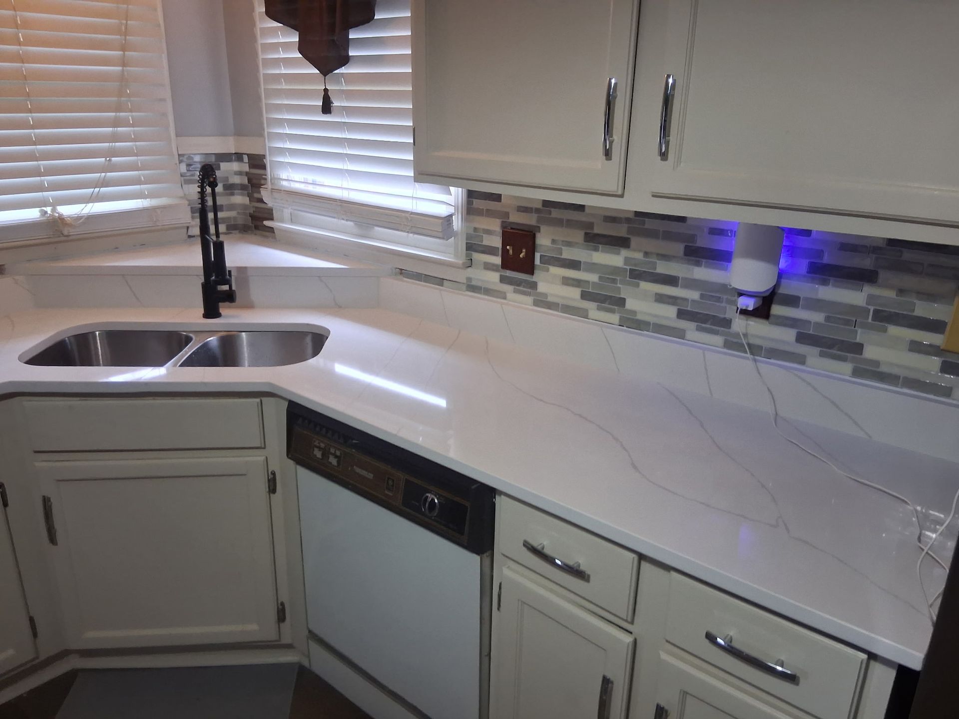 Modern white kitchen with stainless sink, dishwasher, and tile backsplash under cabinet lighting