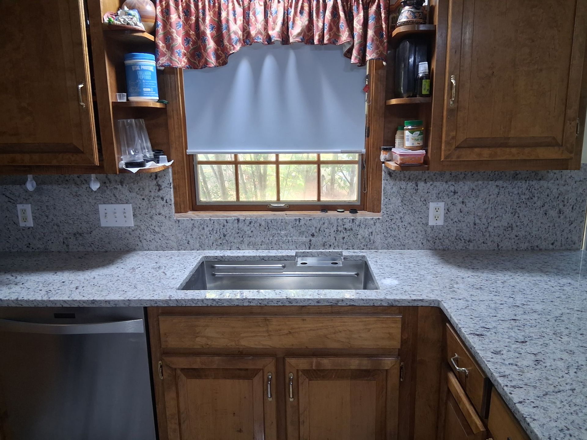 Kitchen sink beneath a window with gray blind, wooden cabinets, and speckled countertop