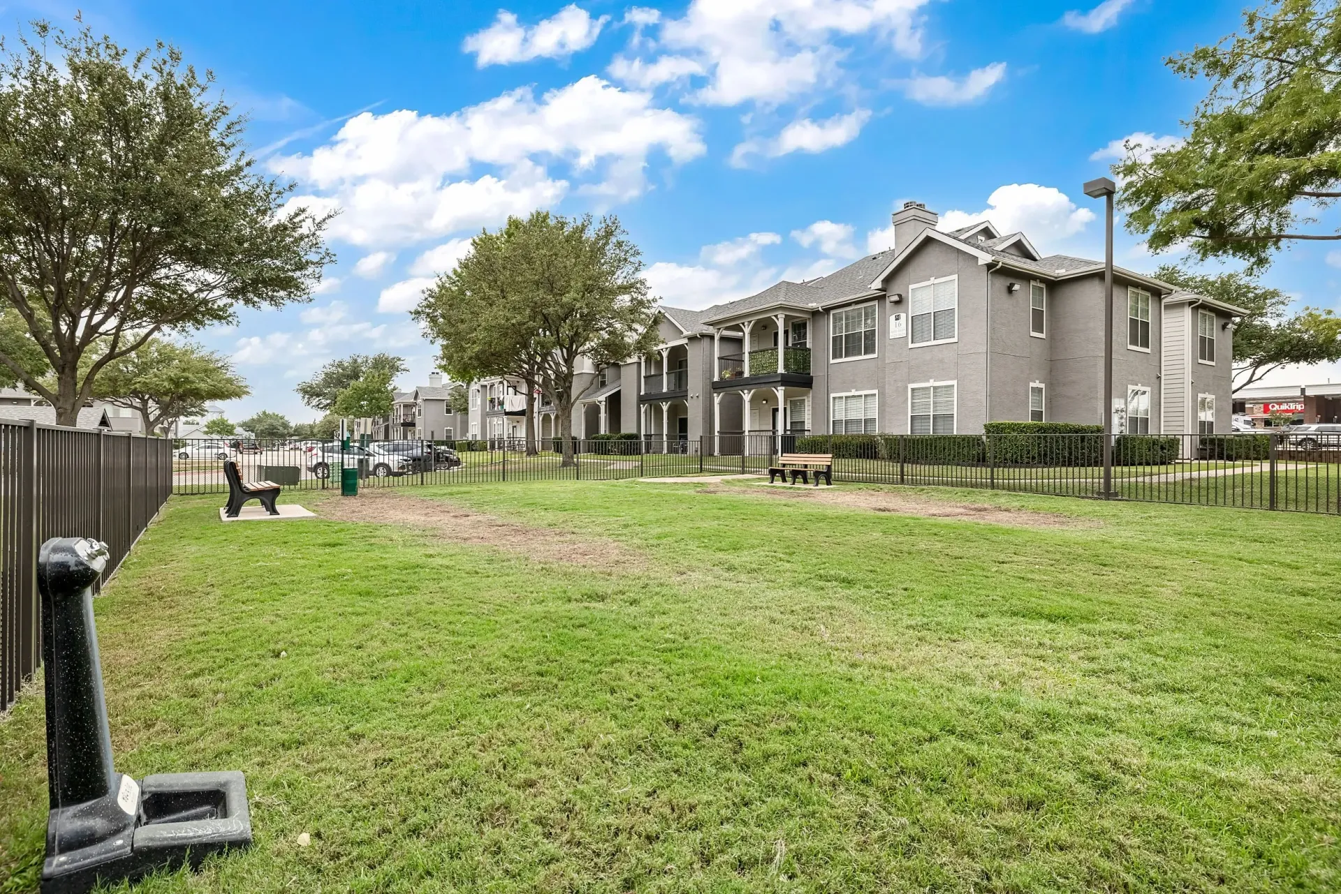 Grassy dog park area with a bench and a dog water station, with apartment buildings in the background.