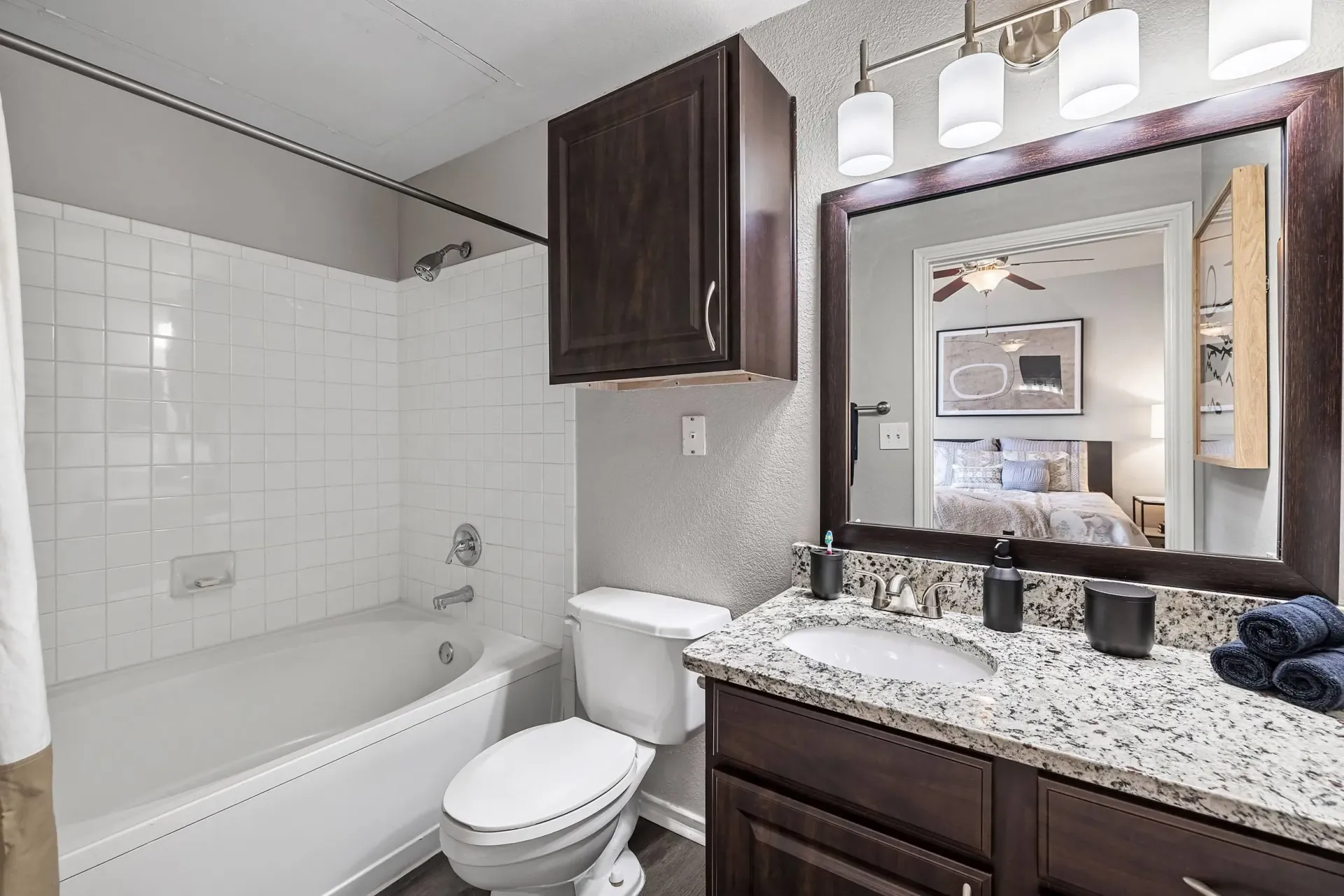Modern bathroom with white tiled shower and tub, granite countertop, and vanity mirror reflecting a bedroom.