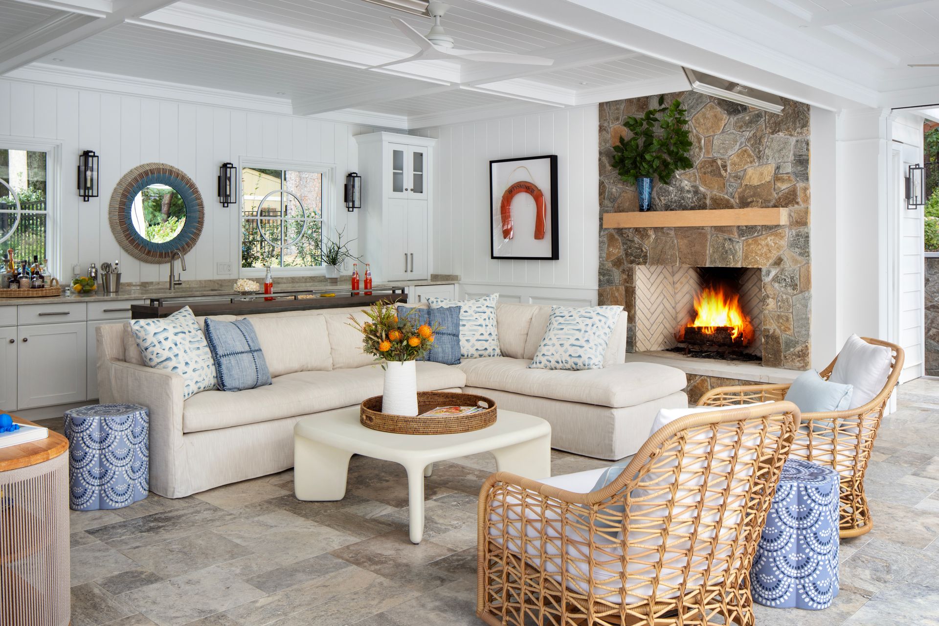 Outdoor living space with fireplace, sofa, and chairs. Stone wall, white cabinets, neutral tones.