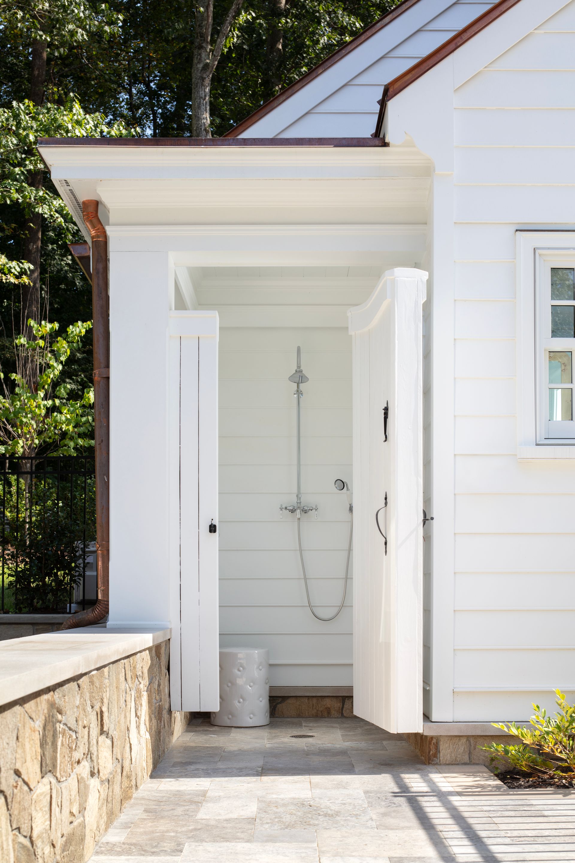 White outdoor shower with open doors, situated by a stone wall, paved path, and white house.
