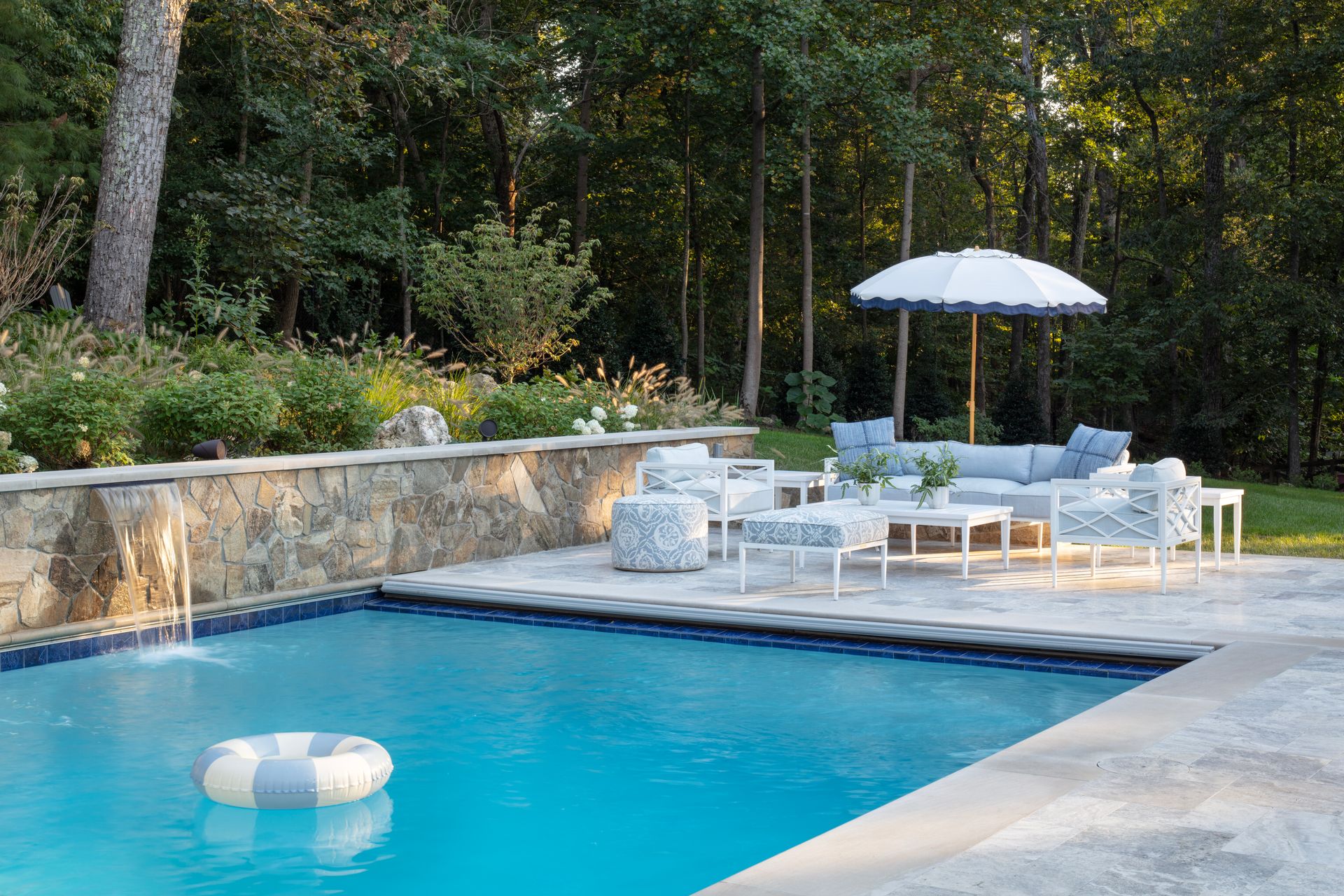 Poolside seating area with blue and white furniture, umbrella, and stone wall with a waterfall.