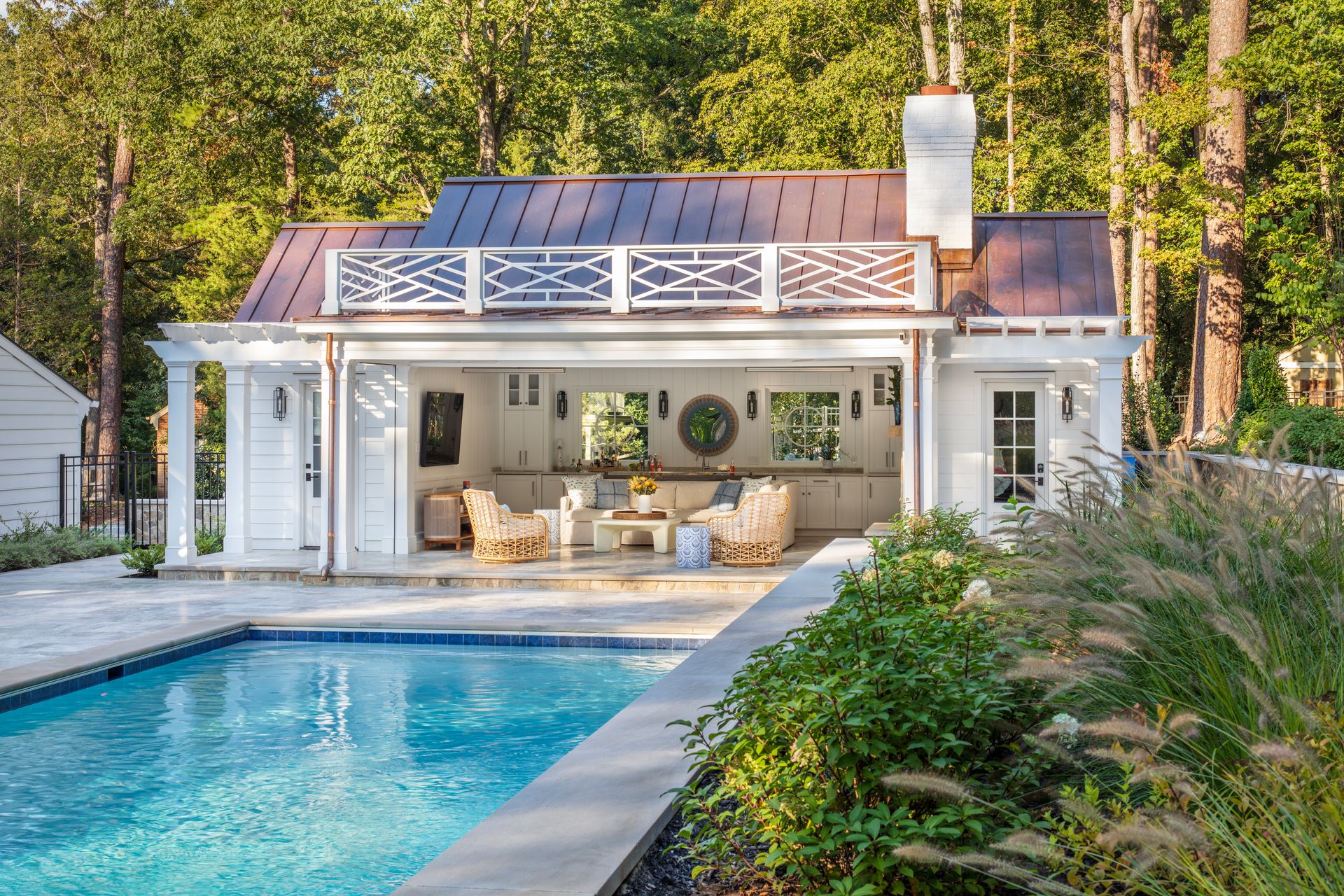 Poolside cabana with open-air kitchen and seating area, copper roof, white exterior, overlooking a blue pool.