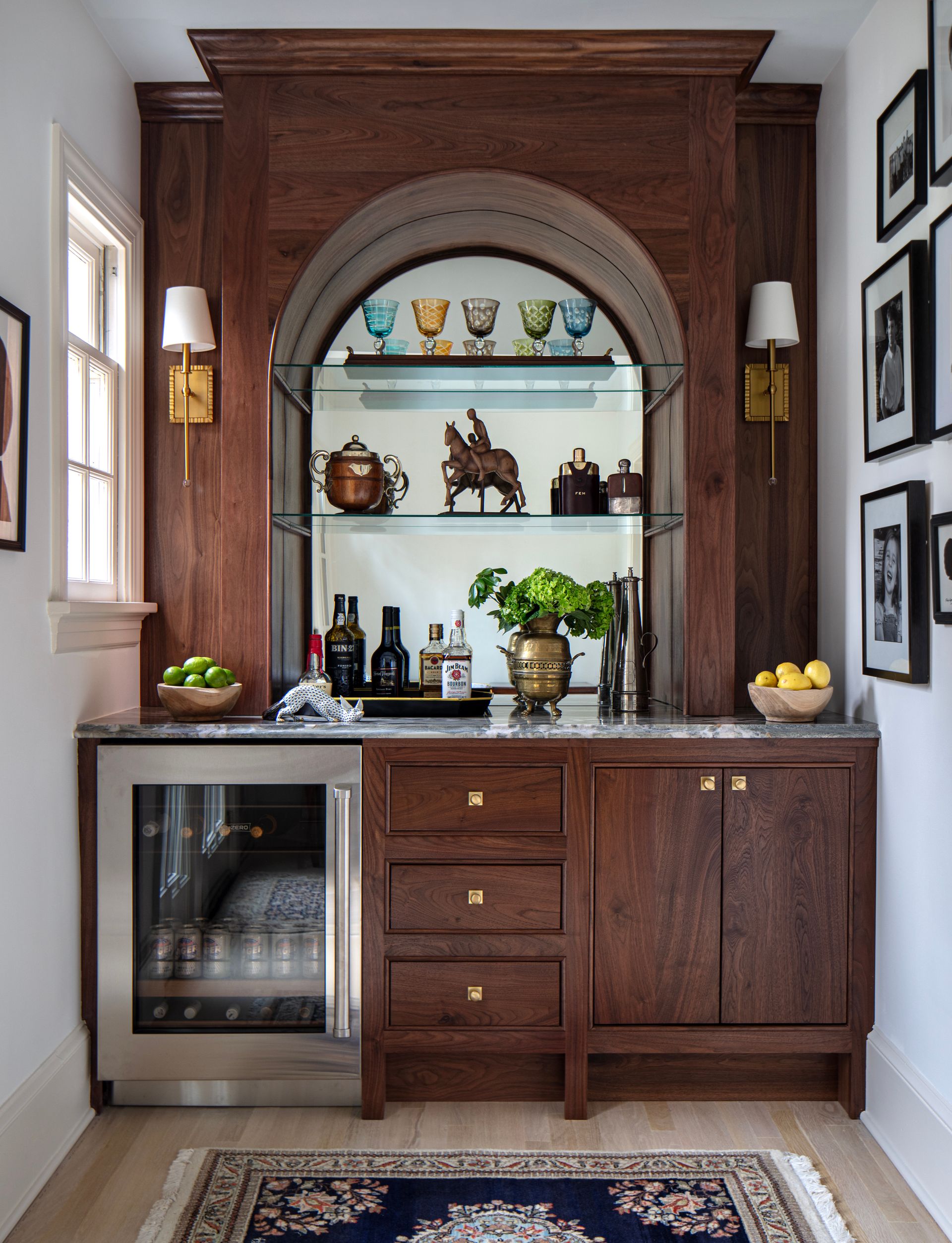 A wooden built-in bar with arched mirror, shelving holding glassware, and mini-fridge.