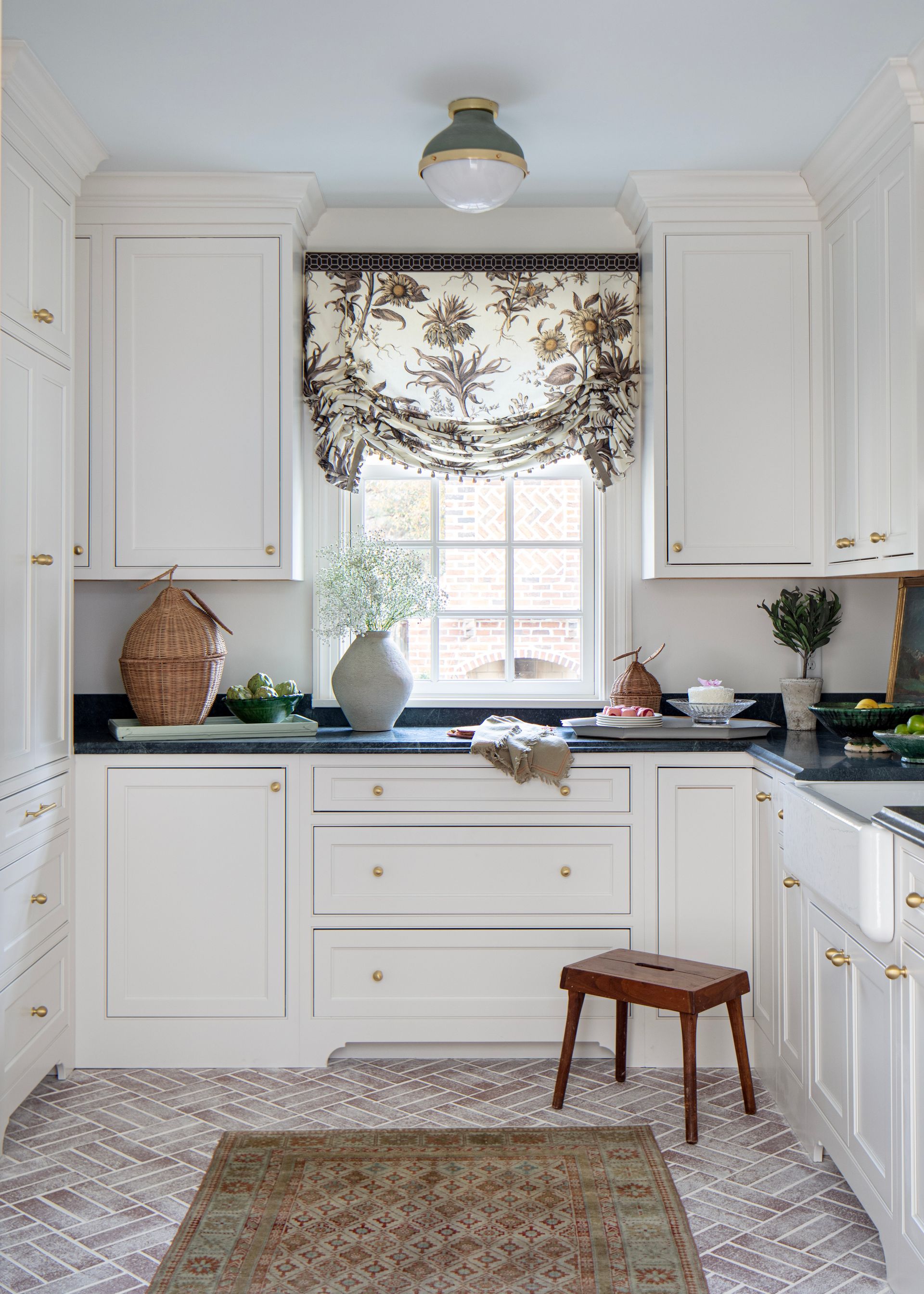 White kitchen with black countertops, floral window shade, patterned floor, and wooden stool.
