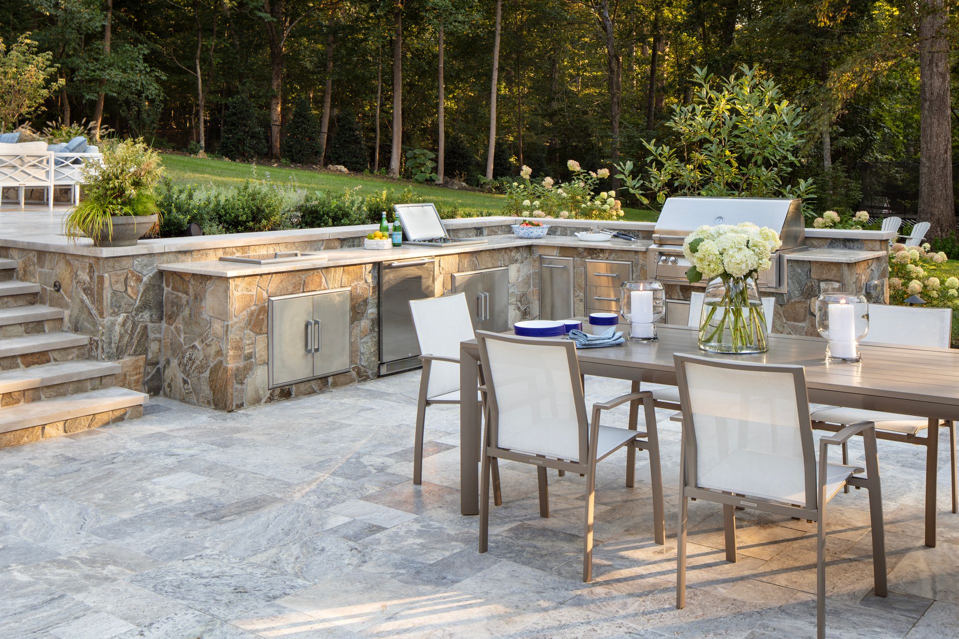 Outdoor patio with built-in kitchen and dining table set for guests. Stone accents, white chairs, and forest backdrop.