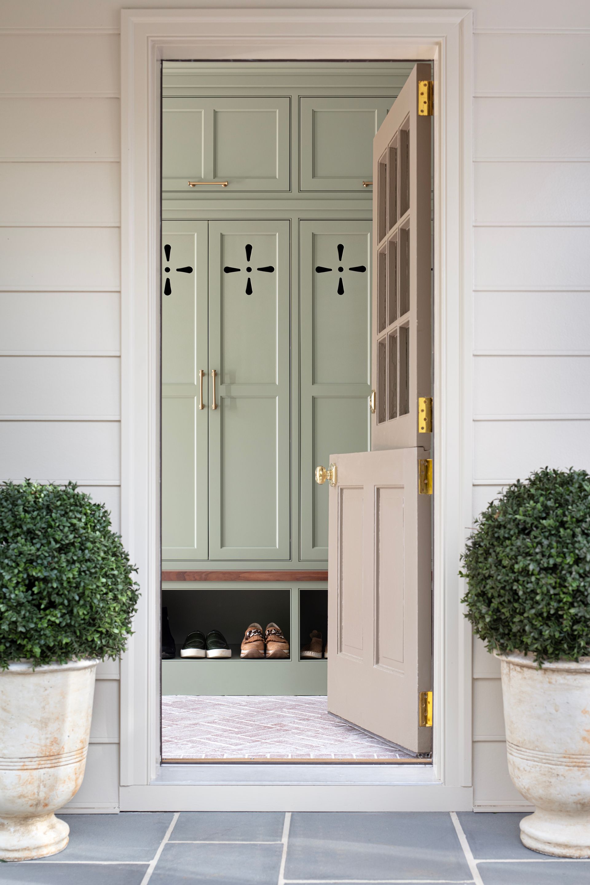 Open, beige door revealing a sage green storage room with shoes on a patterned rug. Two potted bushes flank the doorway.
