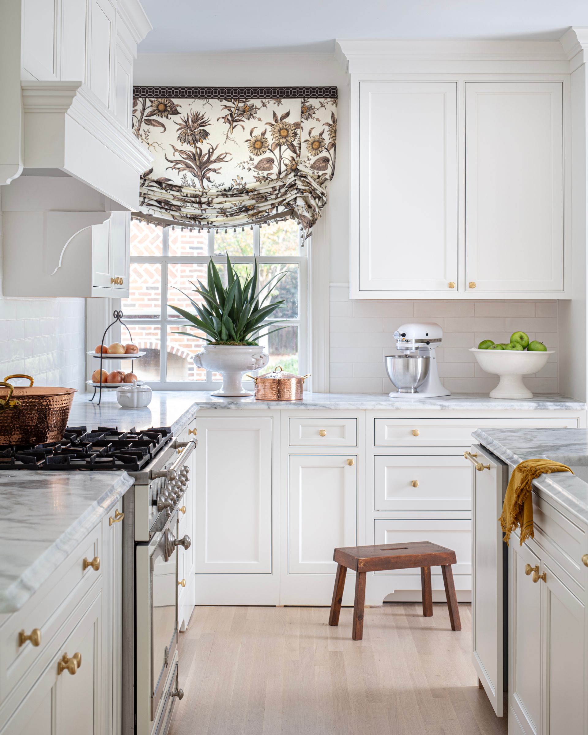 White kitchen with marble countertops, cabinets, and a decorative window shade; a stool sits in the center.
