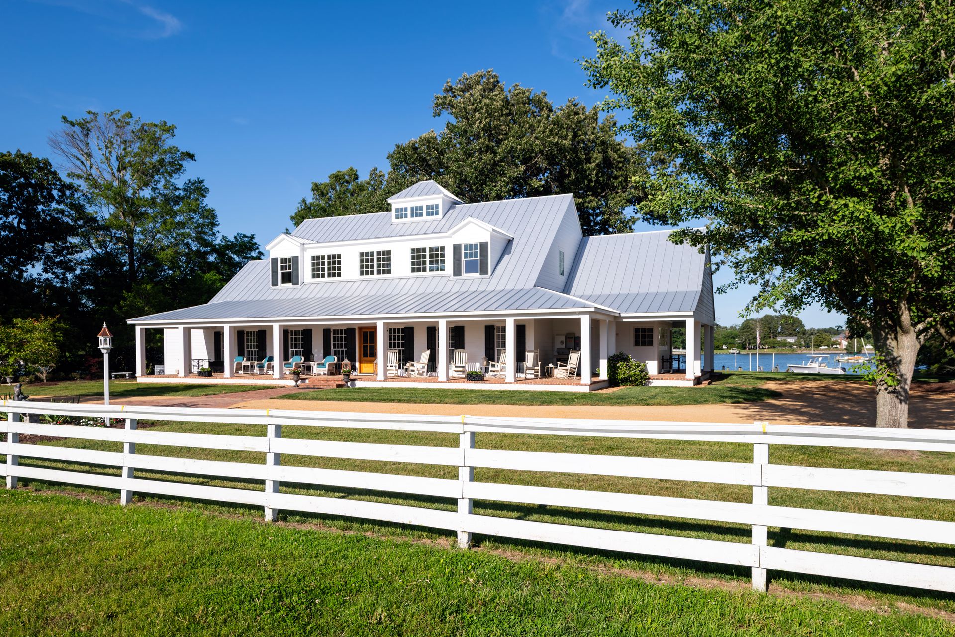 White house with a wraparound porch and a white fence in front; green grass and trees; blue sky.