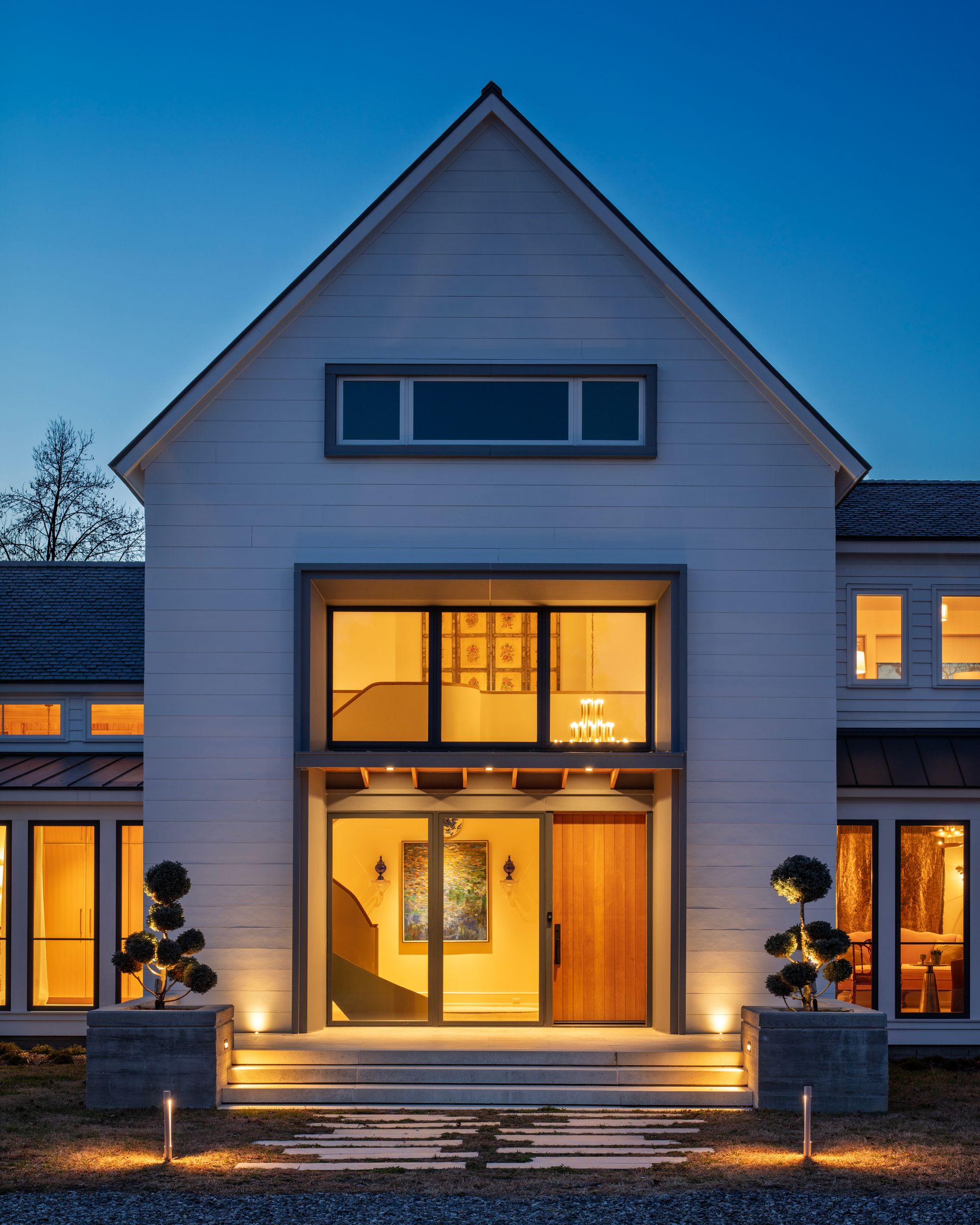 Modern white house with large lit windows at dusk, featuring a glowing front entry and warm interior lighting