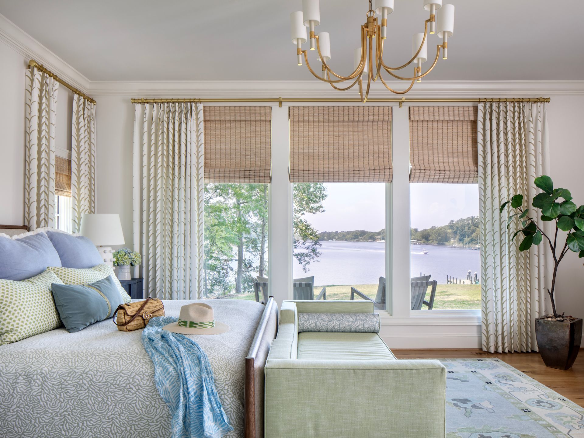 A bright bedroom with a bed facing a large window overlooking a lake.  It features light blue and beige tones, a gold chandelier, and a green potted plant.