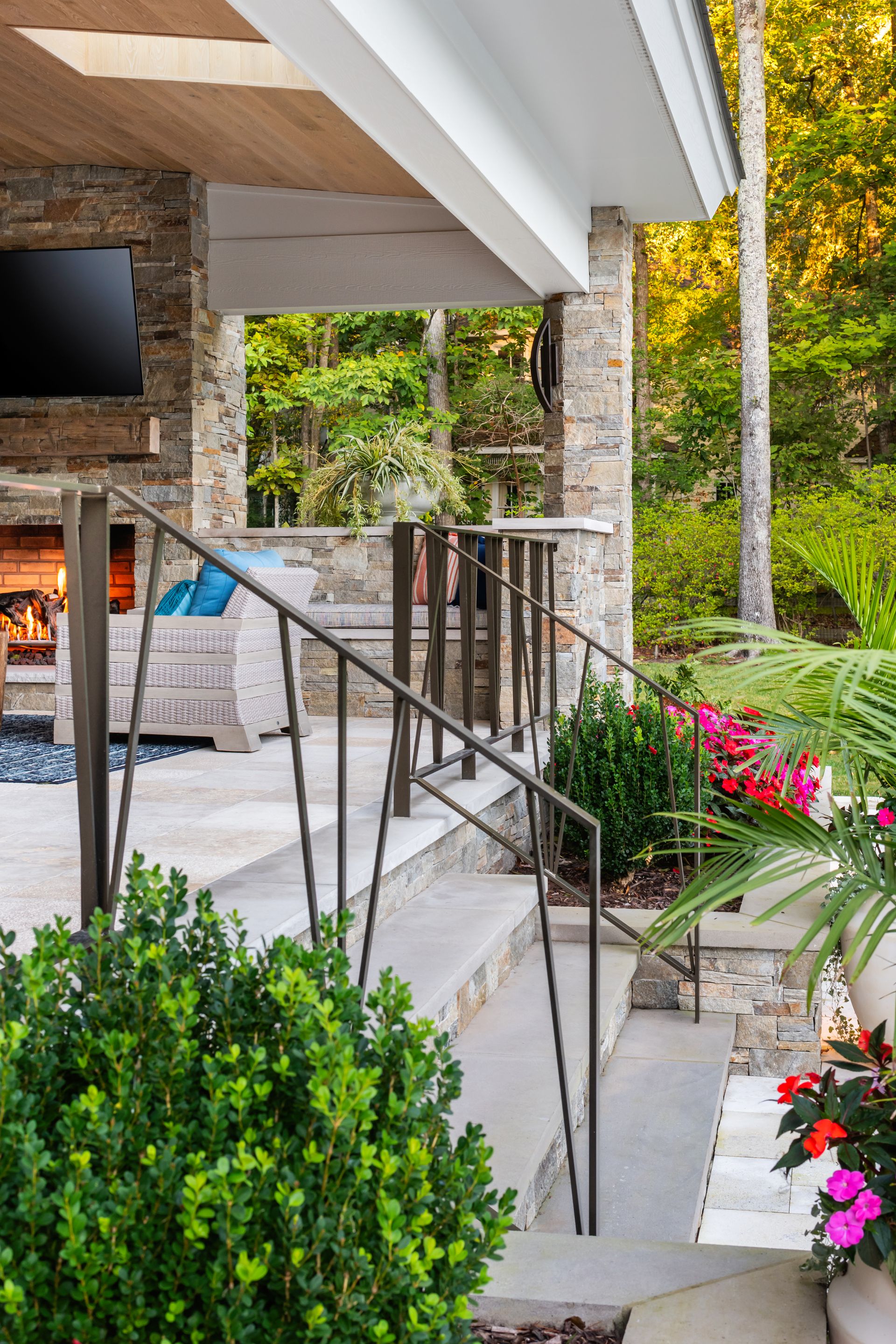Modern patio with stone columns, glass railing, and colorful flowers beside steps and a lush garden.