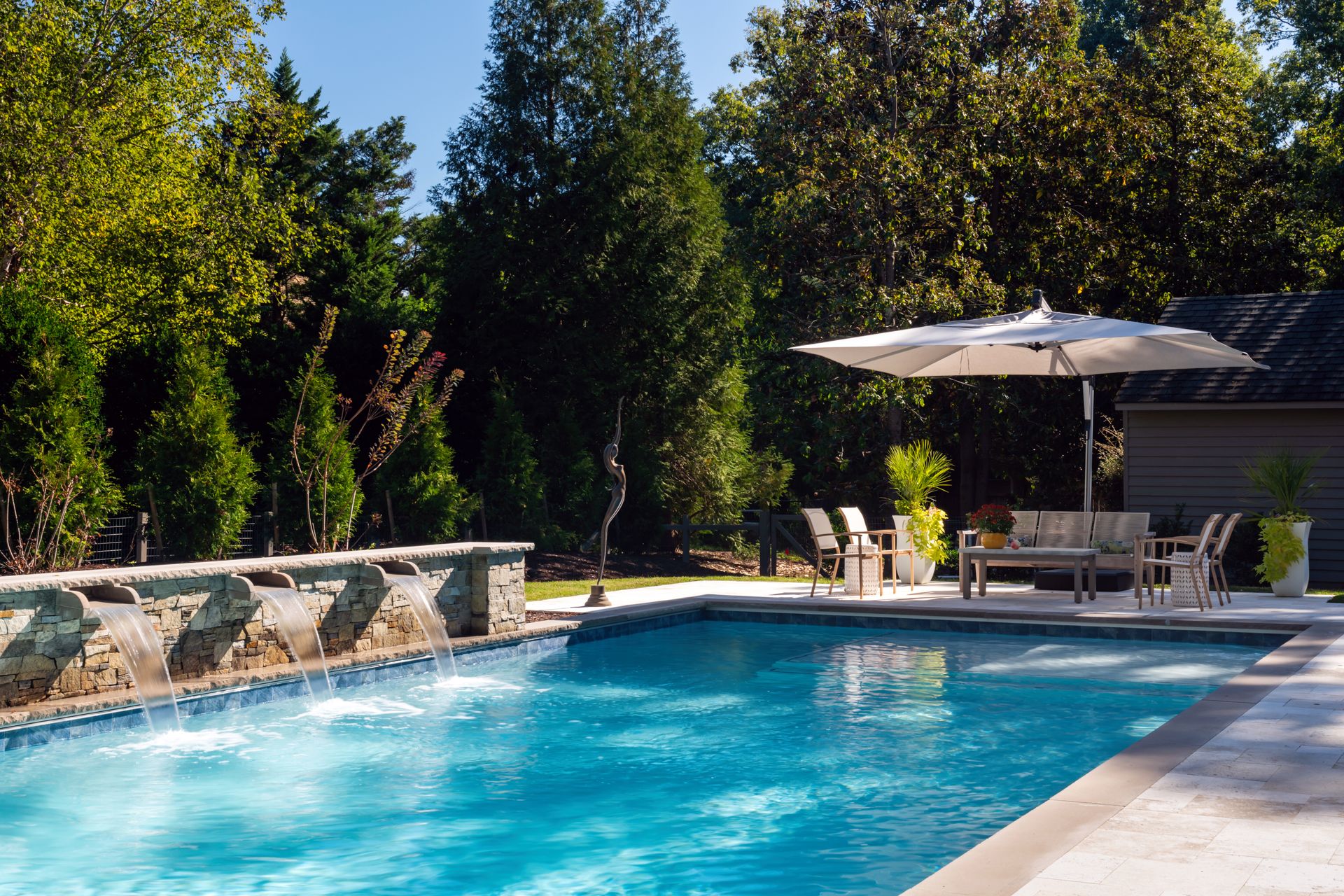 Backyard swimming pool with blue water, stone wall, patio chairs, and a large umbrella under trees