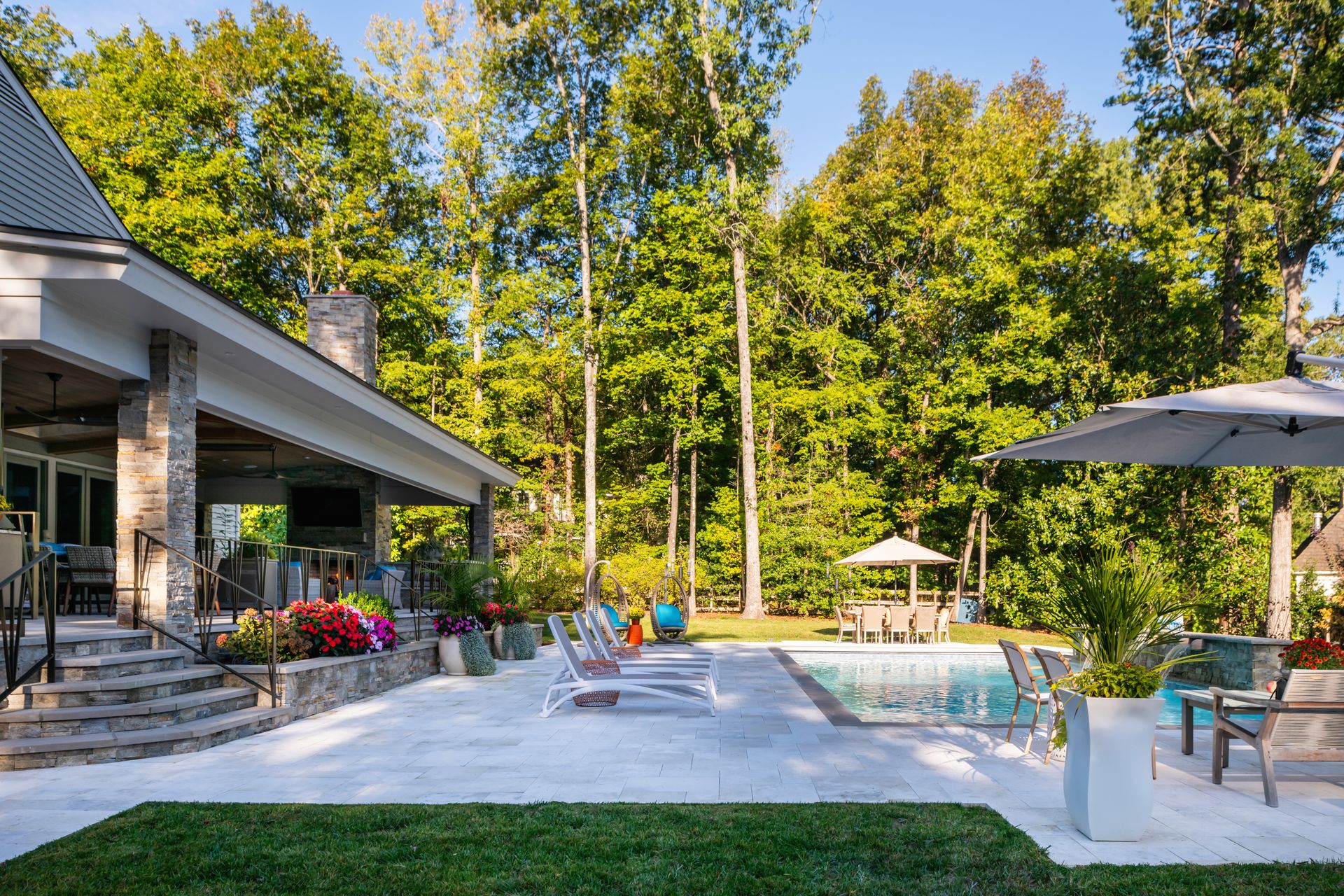 Modern backyard patio with pool, lounge chairs, umbrellas, and a house beside trees