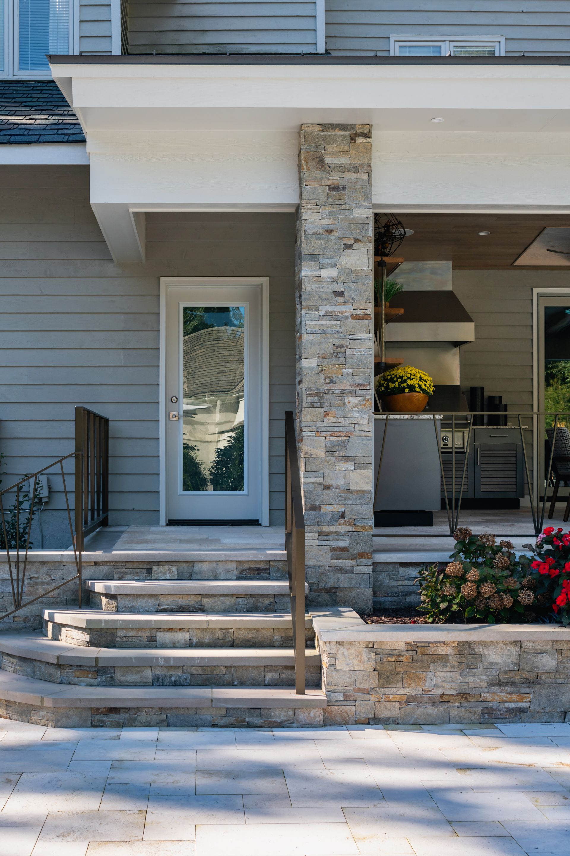 Front porch of a modern house with stone column, steps, glass door, and potted flowers