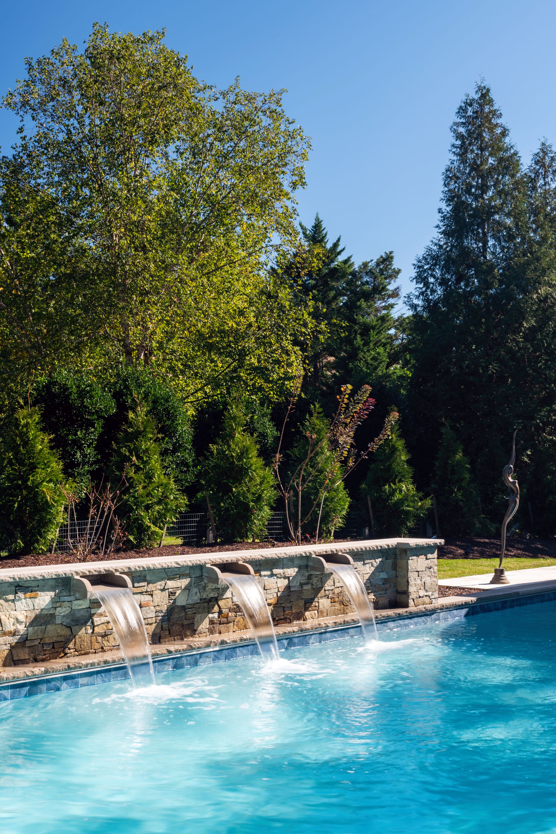 Outdoor pool beside stone wall and trees under a clear blue sky