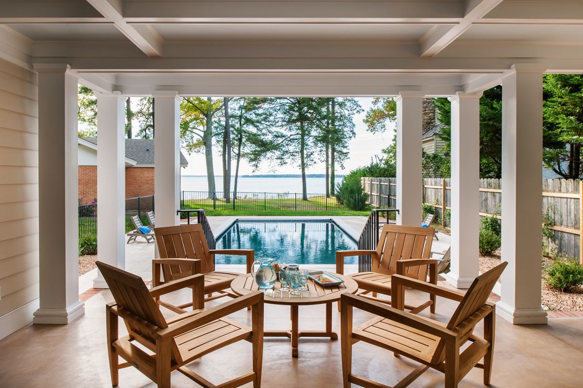 Patio with wooden chairs surrounds a small table, overlooking a pool and a waterfront view with trees.
