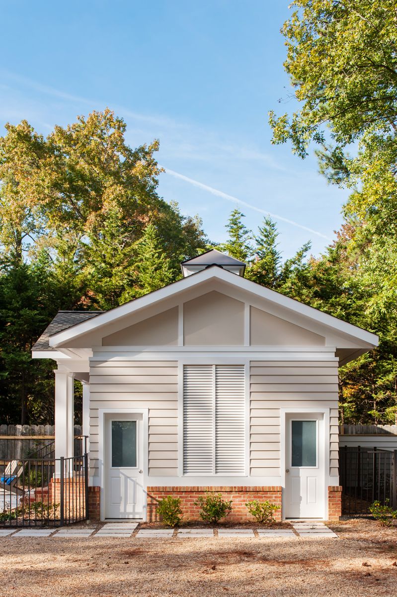 Small, light-colored shed with two white doors, a large central vent, and a peaked roof, set against a backdrop of green trees and blue sky.