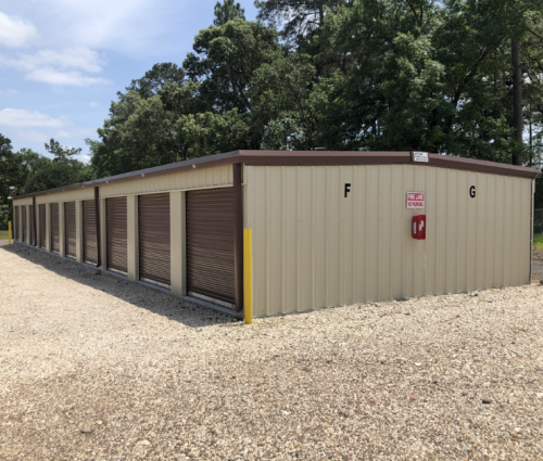 Row of tan storage unit buildings with brown roll-up doors on a gravel lot beside trees.