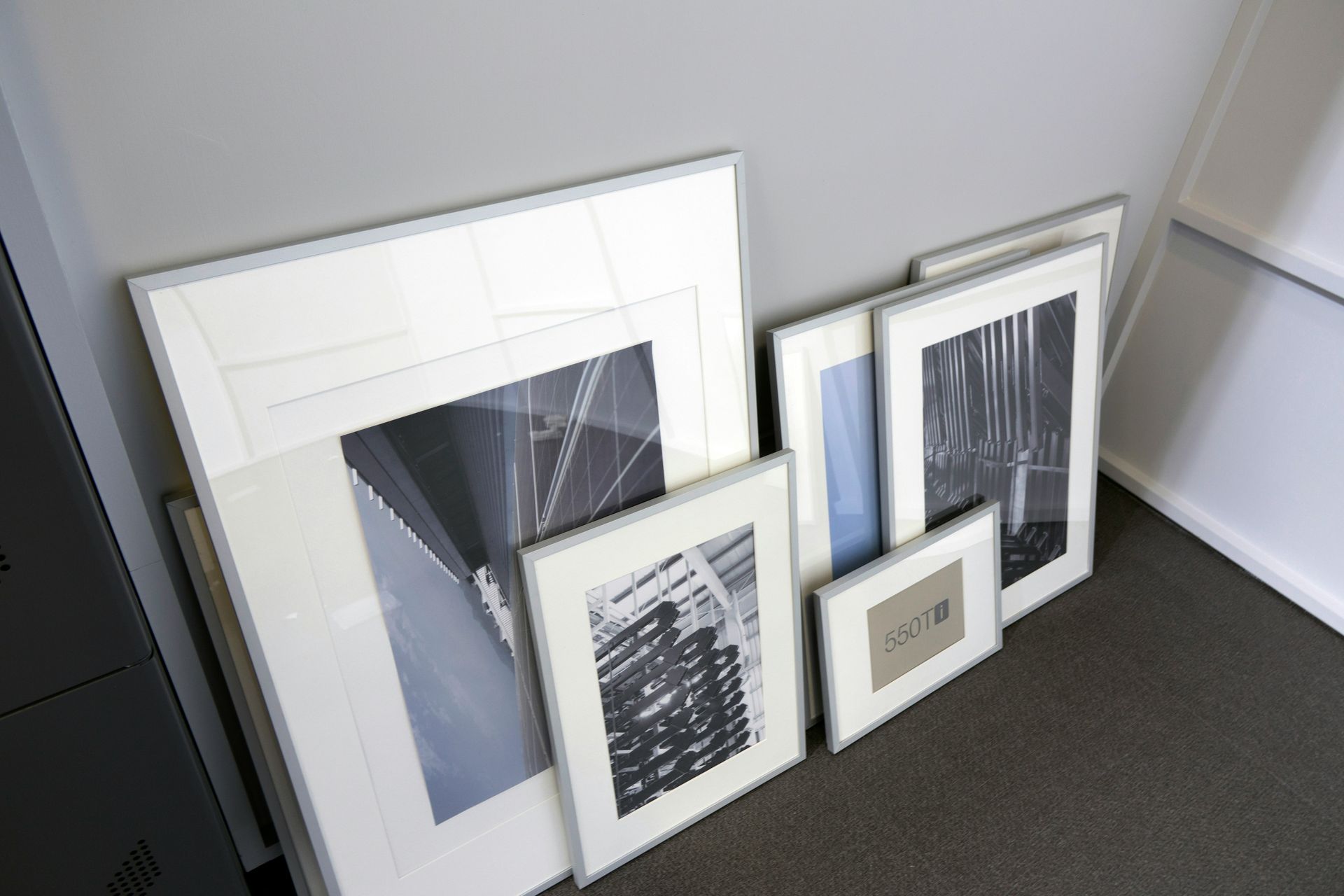 Frames with black and white photos lean against a gray wall on carpet.
