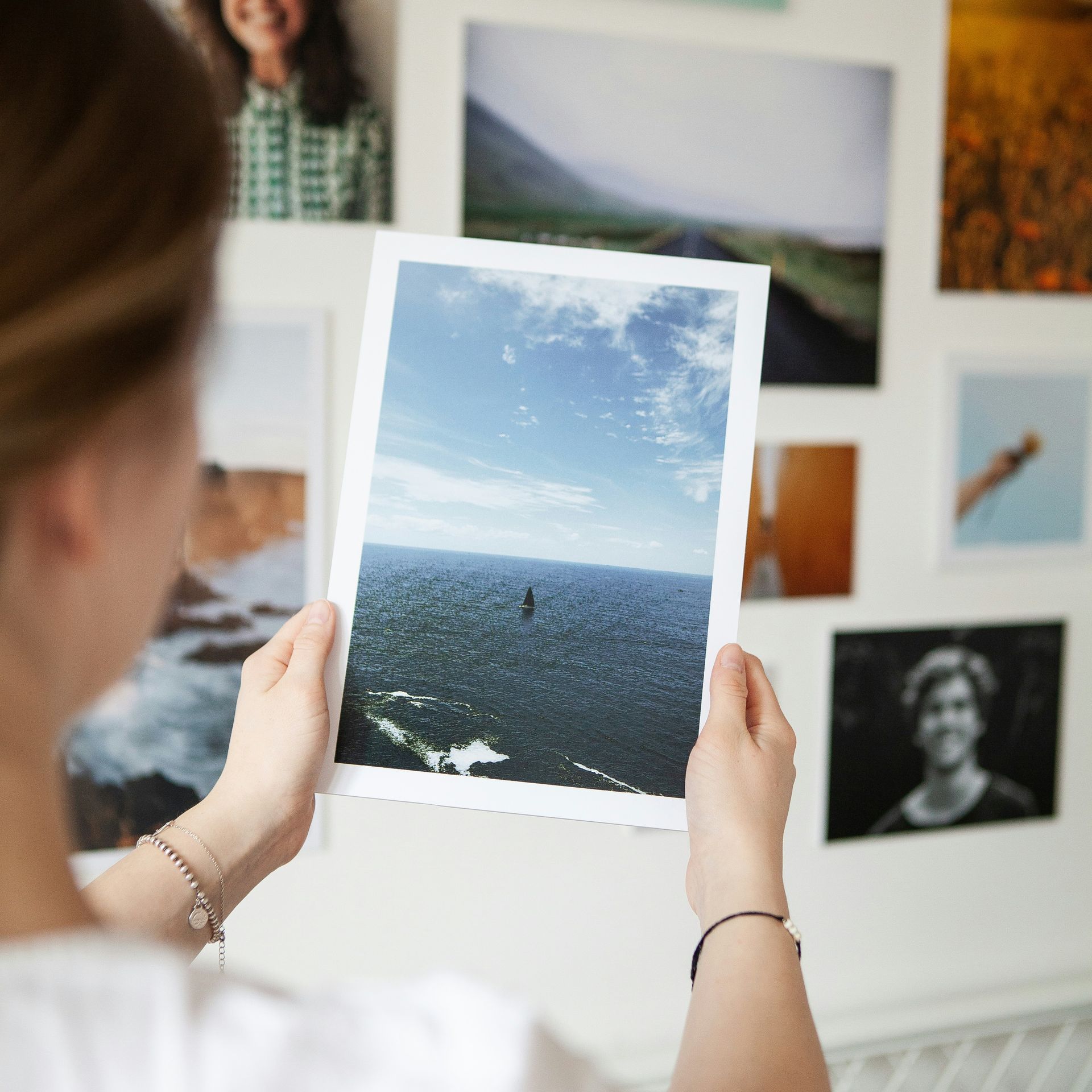 Woman holding a photograph of a blue sky and water, surrounded by other photos on a white wall.