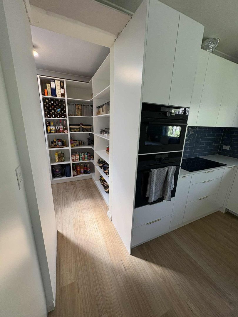 A kitchen featuring a modern white pantry with open shelving, next to a built-in oven unit on a light wood floor.