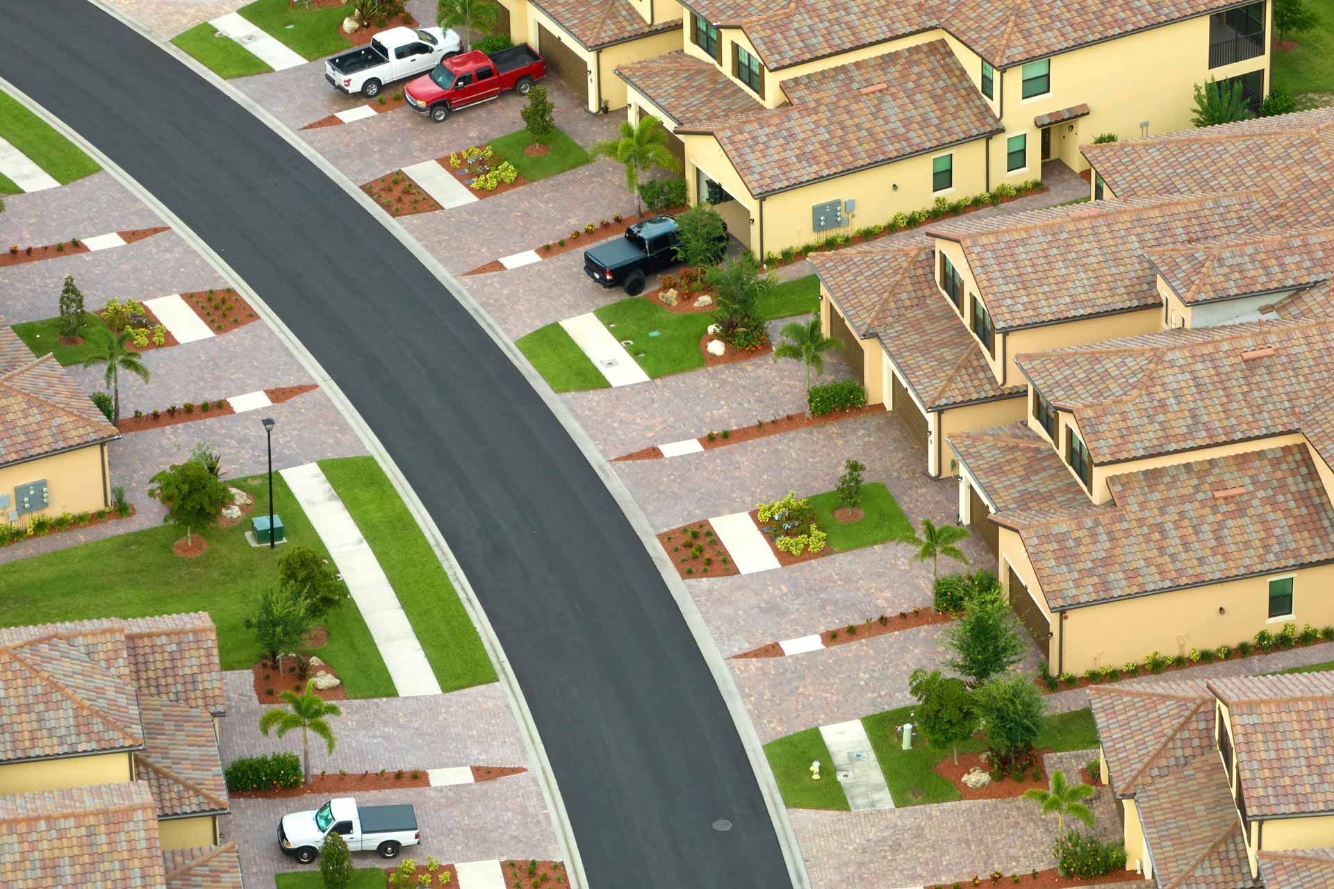Aerial view of a suburban street with attached houses, driveways, green spaces, and parked vehicles.