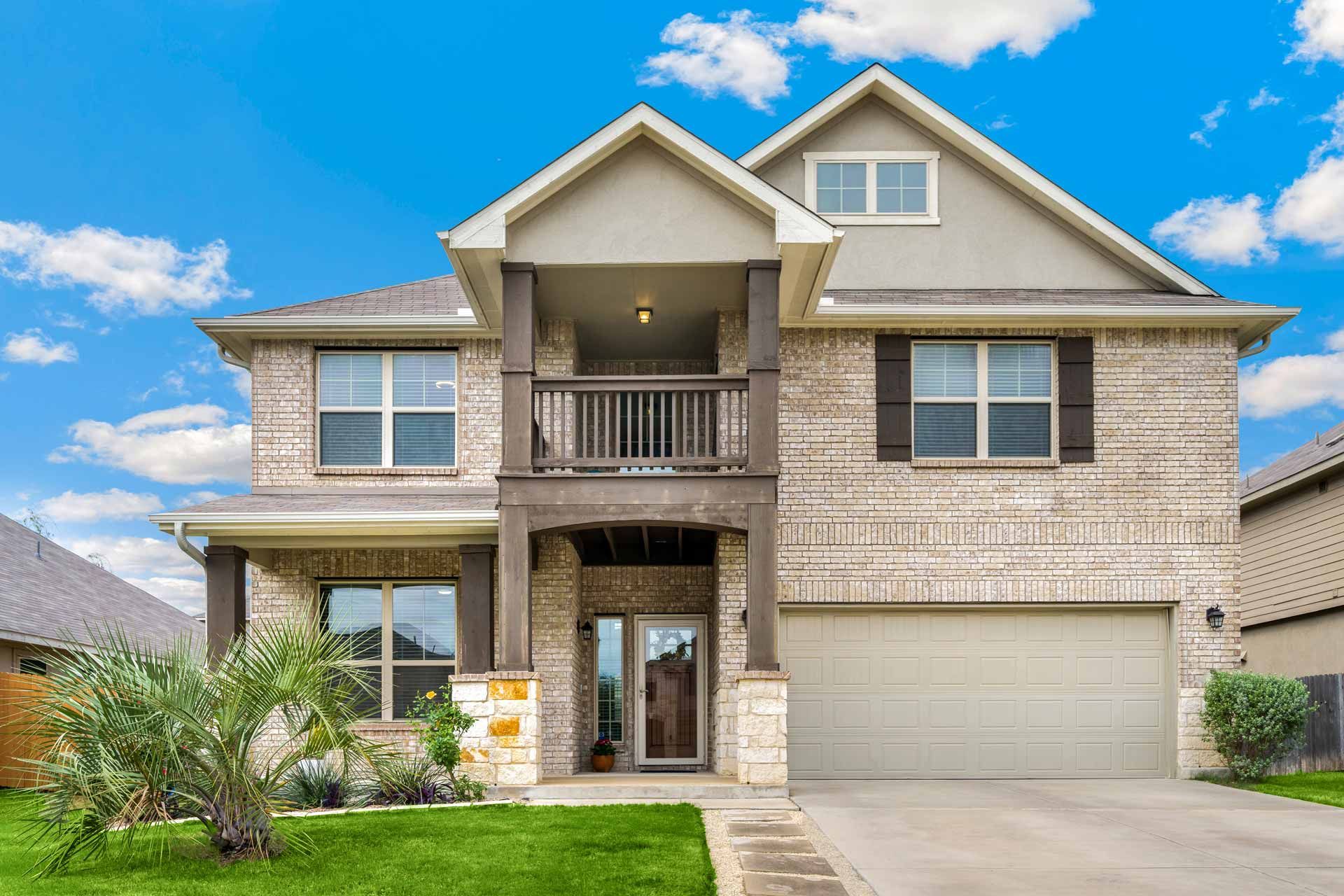 Two-story brick house with a balcony and attached garage, under a blue sky.