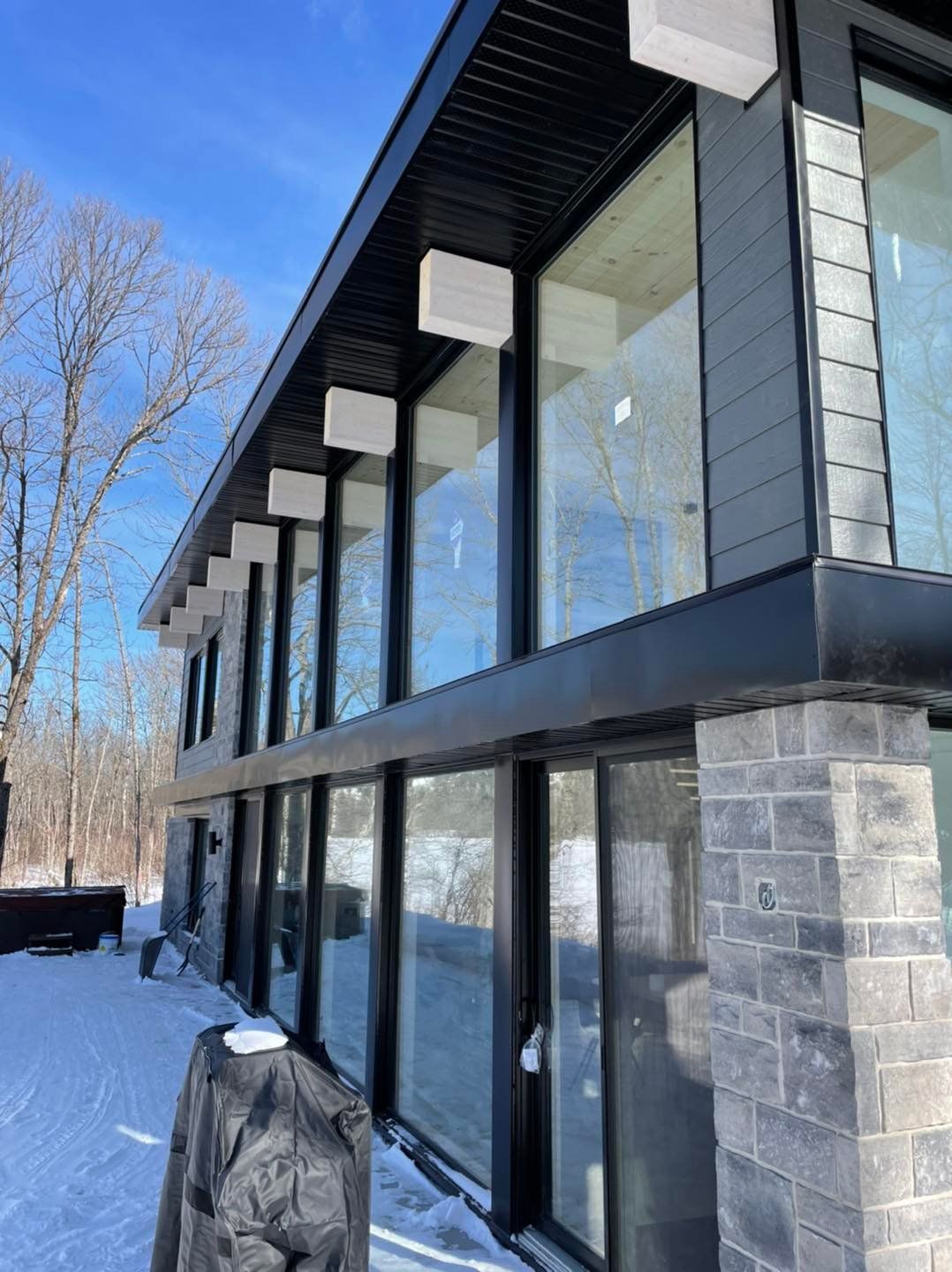 Maison moderne à deux étages avec de grandes fenêtres, des piliers en pierre et des touches de noir dans un décor extérieur enneigé.