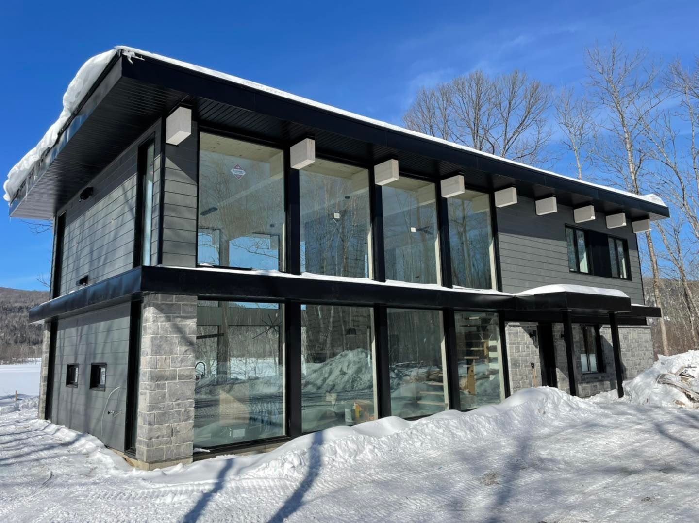 Maison moderne à deux étages avec de grandes fenêtres et un toit plat, entourée de neige sous un ciel bleu.